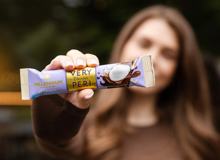 Жінка з довгим каштановим волоссям тримає шоколадний батончик Millennium Very Peri Coconut у напрямку до камери, при цьому упаковка та зображення кокоса знаходяться у фокусі, а її обличчя розмите на задньому плані.