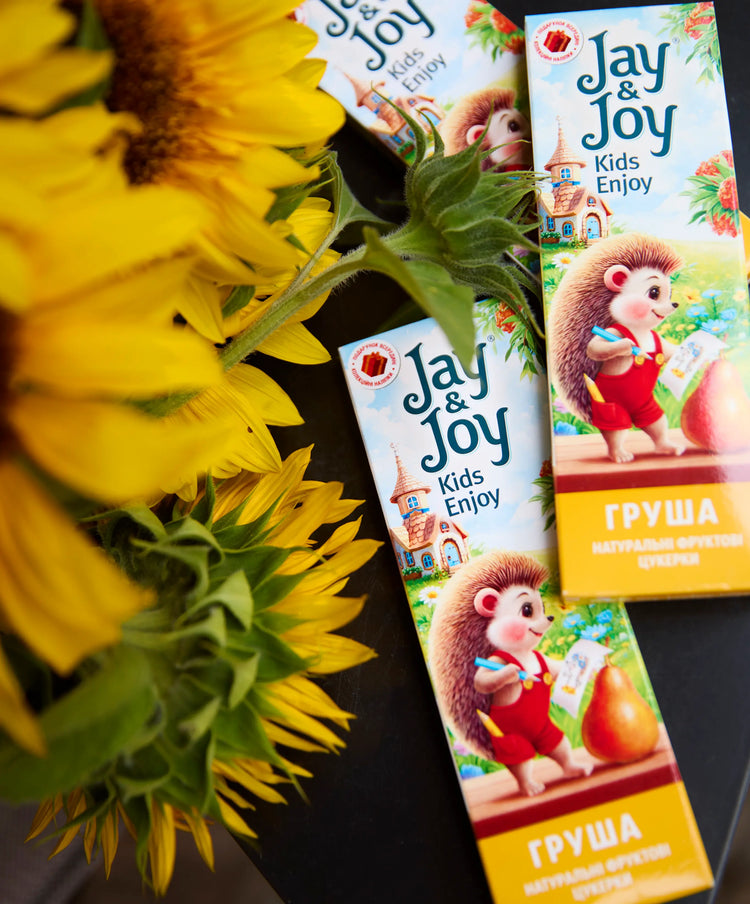 Дві барвисті коробки фруктових снеків Jay & Joy Kids Enjoy з мультяшними їжачком та грушею розміщені поруч з яскравими жовтими соняшниками на темній поверхні.