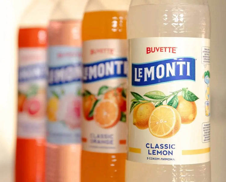 Чотири пляшки напоїв Buvette Le Monti з різними смаками, з акцентом на пляшці Classic Lemon із зображенням лимона; інші смаки розмиті на задньому плані.