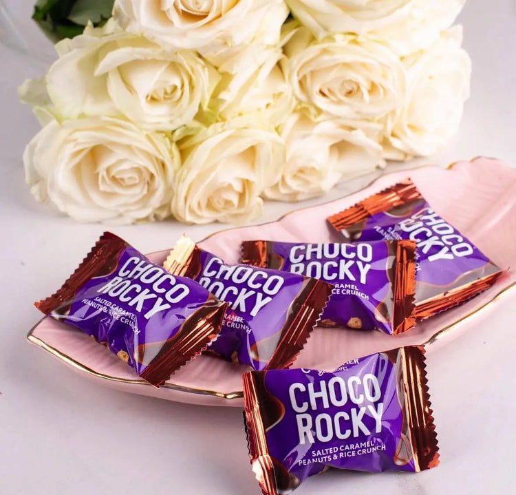 Рожева тарілка з кількома цукерками Choco Rocky у фіолетовій обгортці стоїть поруч із букетом білих троянд на білій поверхні.