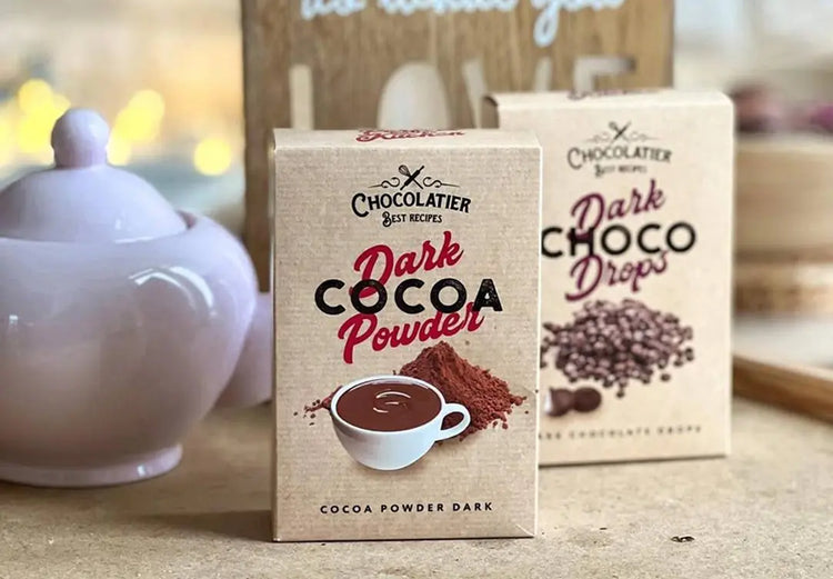 Дві коробки какао-продуктів бренду Chocolatier, одна з яких - "Темний какао-порошок" із зображенням какао-порошку та чашки, а інша - "Темні шоколадні краплі", розміщені на дерев'яній поверхні.