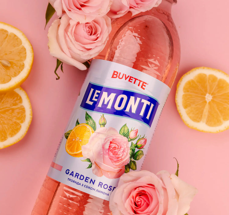 Рожева пляшка напою Le Monti Garden Rose лежить на рожевій поверхні в оточенні розрізаних навпіл лимонів і рожевих троянд. На етикетці - зображення троянд і лимонів.