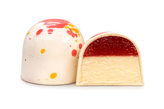 Кондитерський виріб у формі купола з білого шоколаду — «Millennium Craft Strawberry & Lemon with Stevia» від Millennium Chocolate — прикрашений червоними та жовтими бризками; на фото він зображений цілим та розрізаним навпіл, що дозволяє побачити шар полуничного мармеладу поверх кремової білої начинки.