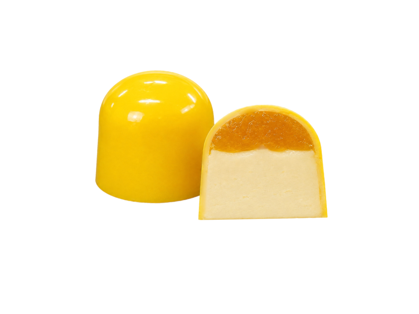 Дві жовті цукерки у формі купола «Millennium Craft Mango & Coconut with Stevia» від Millennium Chocolate виготовлені з білого шоколаду. Одна з них ціла, а інша розрізана навпіл, щоб було видно кремово-білу начинку з кокосового пюре, покриту яскравим шаром мангового мармеладу. Обидві цукерки розміщені на однотонному білому тлі.
