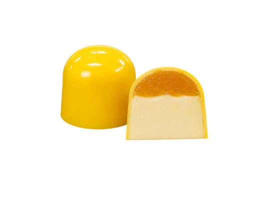Дві жовті цукерки у формі купола «Millennium Craft Mango & Coconut with Stevia» від Millennium Chocolate виготовлені з білого шоколаду. Одна з них ціла, а інша розрізана навпіл, щоб було видно кремово-білу начинку з кокосового пюре, покриту яскравим шаром мангового мармеладу. Обидві цукерки розміщені на однотонному білому тлі.